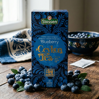 Opakowanie Stassen Blueberry Ceylon Tea 25 torebek, otoczone świeżymi borówkami na drewnianym stole przy oknie.